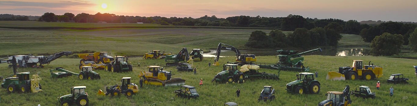 Cilvēki pastaigājas starp vairāku veidu John Deere mašīnām lielā laukā.