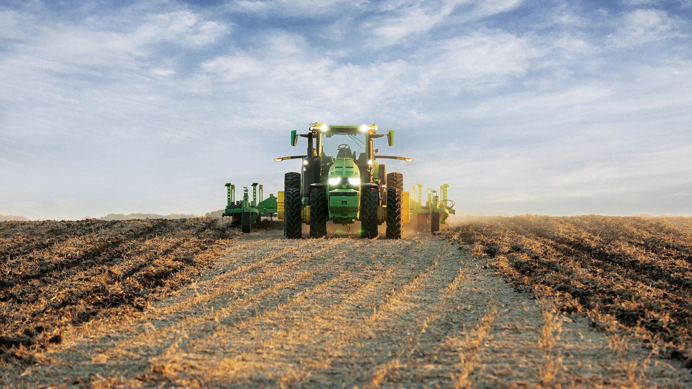 John Deere traktora pašvadāmība, velkot zemes apstrādes aprīkojumu pa atvērtu lauku.
