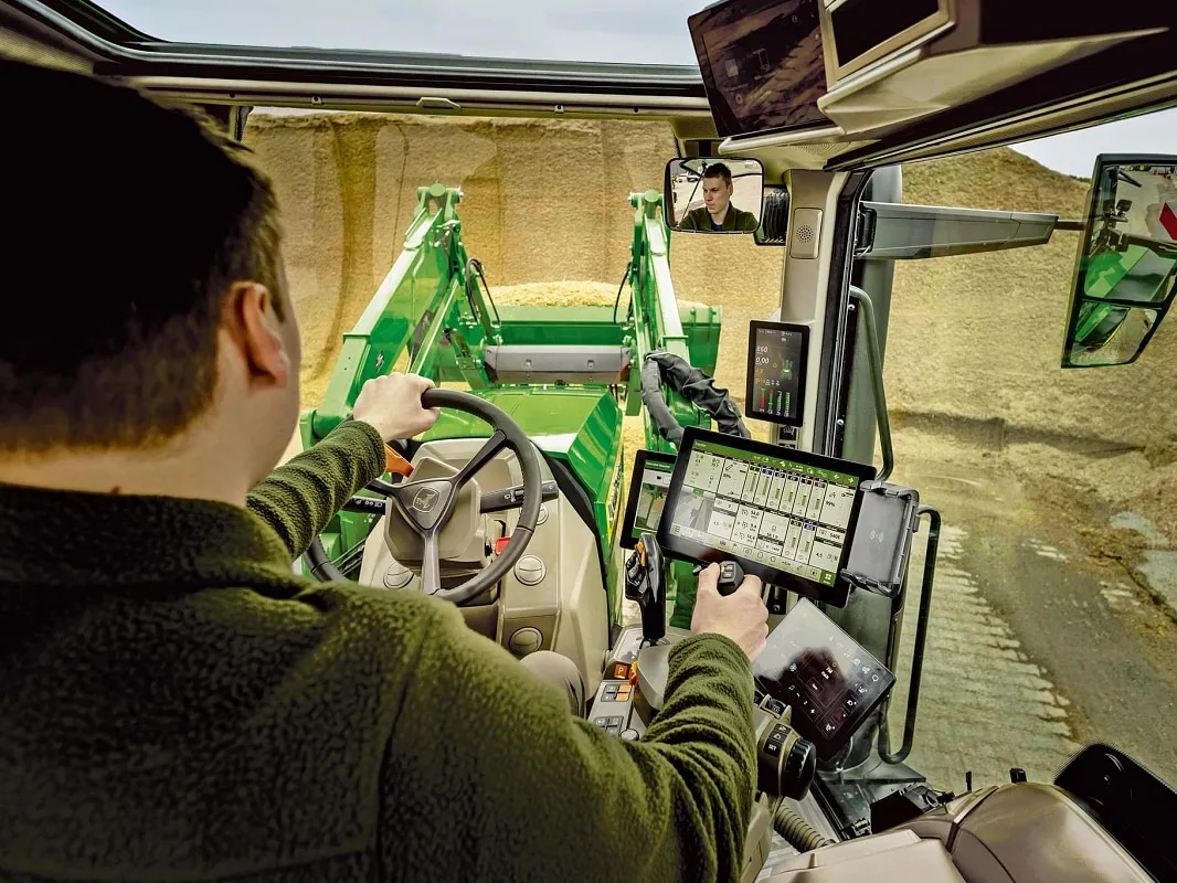 John Deere traktora kabīnes iekšpuses skats, kurā redzamas operatora vadības ierīces un frontālā iekrāvēja darbība.