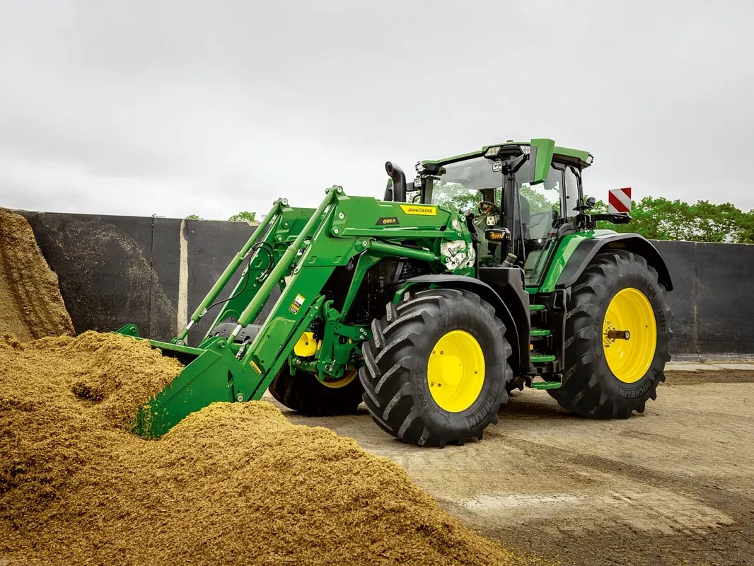 John Deere traktors ar priekšējā iekrāvēja kausu stumj skābbarības kaudzi saimniecības vidē.