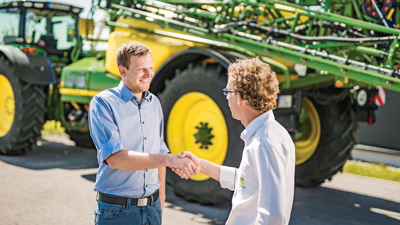 Izplatītāja un klienta rokasspiediens ar John Deere miglotāju fonā