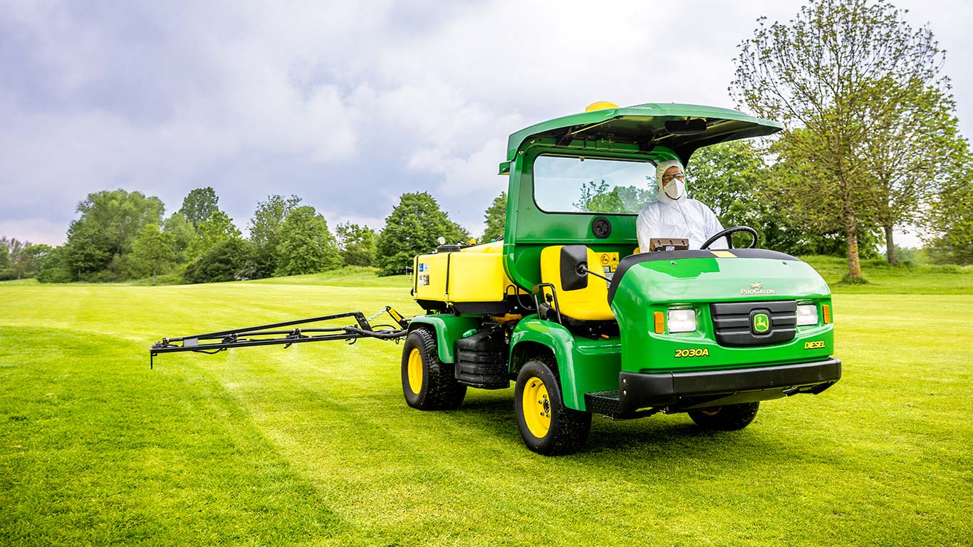 Golfa laukumu zālienu apsaimniekotājs aizsargapģērbā strādā golfa laukuma zālājā ar John Deere smidzinātāju.