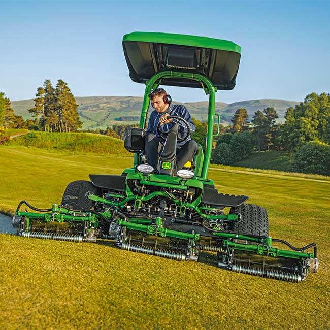 Golfa laukumu zālienu apsaimniekotājs strādā paklājzāliena zonā ar John Deere braucamo grīna pļāvēju ar skatu no priekšpuses.