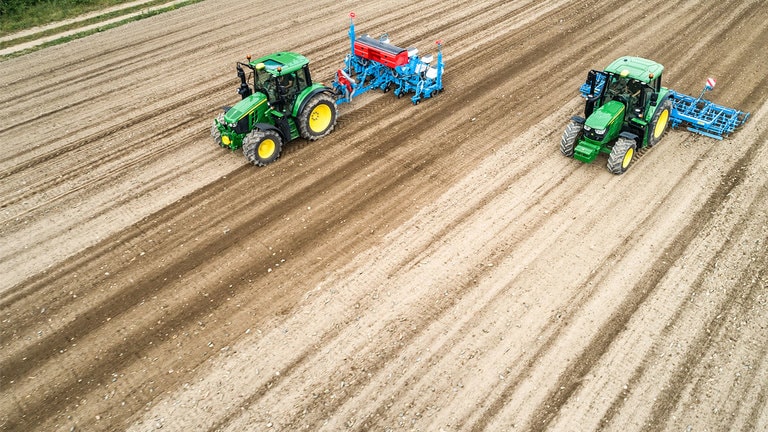 Divi John Deere 6M traktori uz neapstrādātas zemes lauka. Viens traktors velk Monsomen stādītāju.  Otrs traktors velk Lemken sēklu gultnes veido&scaron;anas agregātu
