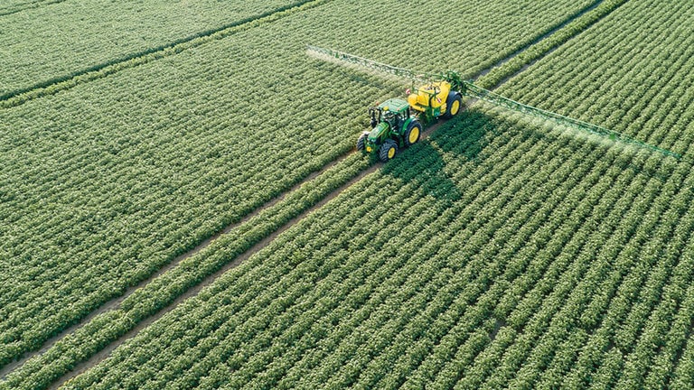 John Deere 6M sērijas traktors velk M700 piekabināmo smidzinātāju kartupeļu laukā