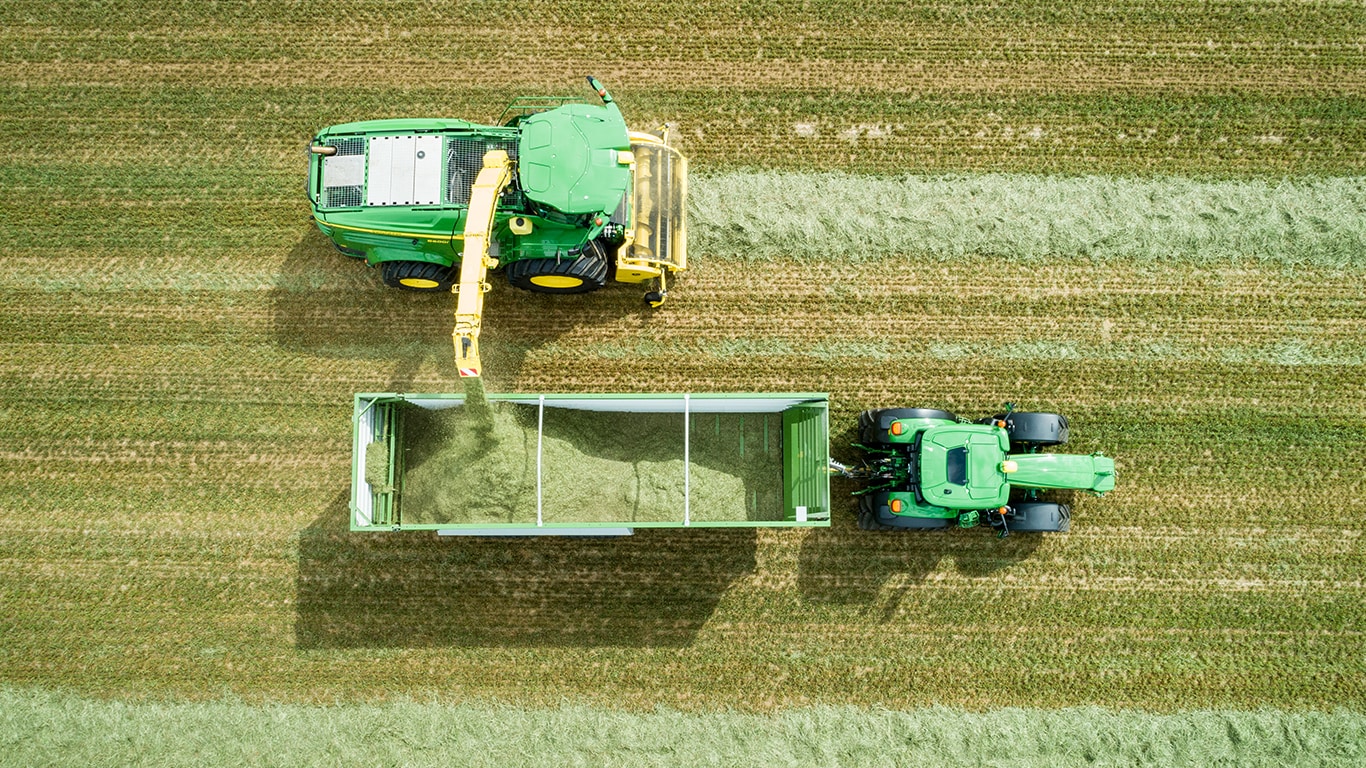 Aerofotouzņēmums ar John Deere 8000 sērijas pa&scaron;gājēju kombainu ar zāles savācēju, kas skābbarību iekrauj piekabē, ko velk John Deere traktors