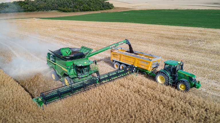 John Deere X sērijas kombains veic rap&scaron;u izkrau&scaron;anu graudu ratiņos, ko velk John Deere traktors