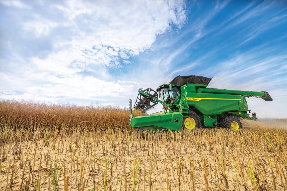 John Deere kombains novāc graudus ar hederu laukā zem zilām debesīm ražas novākšanas laikā