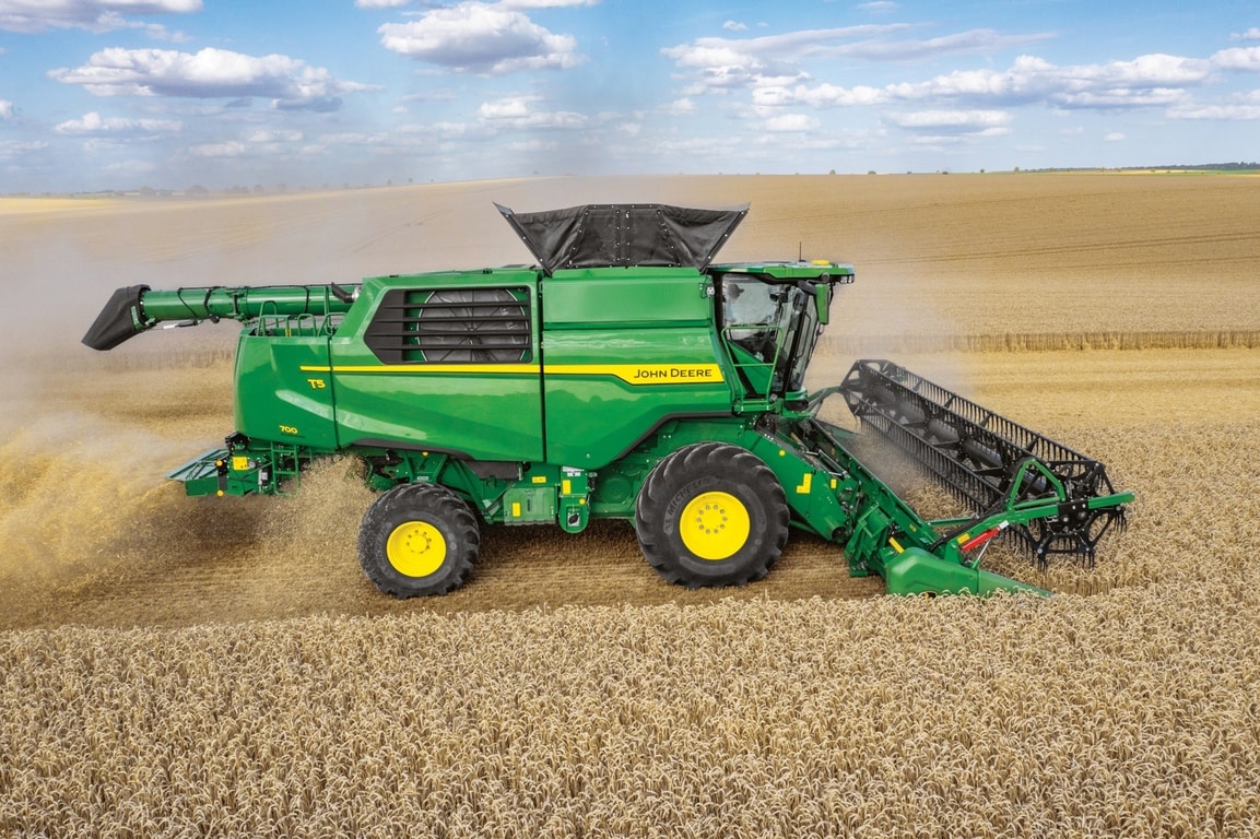 John Deere kombaina sānskats ar pļaušanas hederu, kas novāc kviešus lielā lauksaimniecības laukā