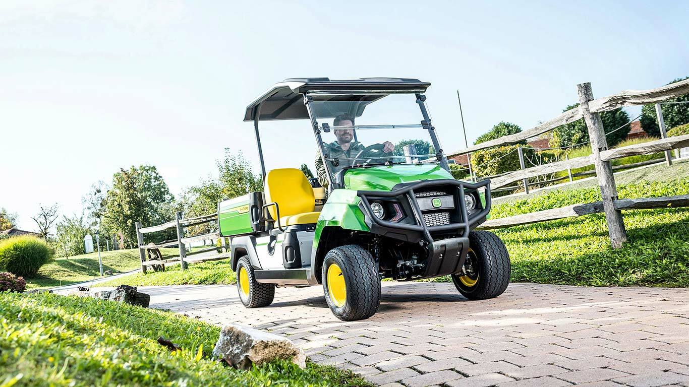 Zaļš John Deere Gator GS universālais transportlīdzeklis brauc pa asfaltētu celiņu cauri labiekārtotai teritorijai ar zāli un koka žogiem.