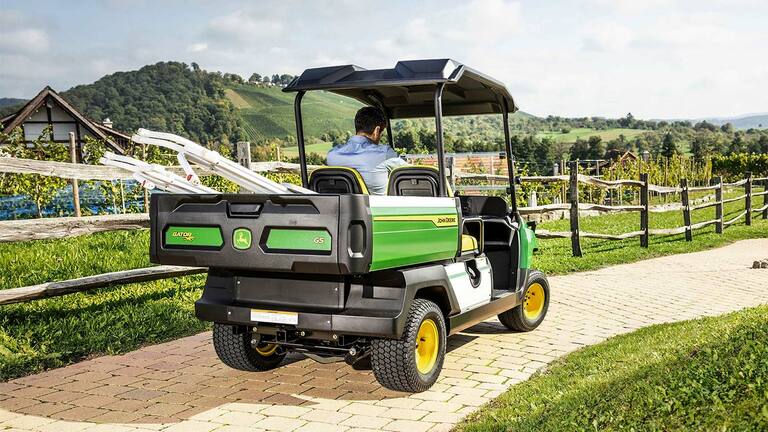 Atpakaļskats uz John Deere Gator GS, braucot pa ķieģeļu taku ar gleznainiem pakalniem un vīna dārziem fonā.