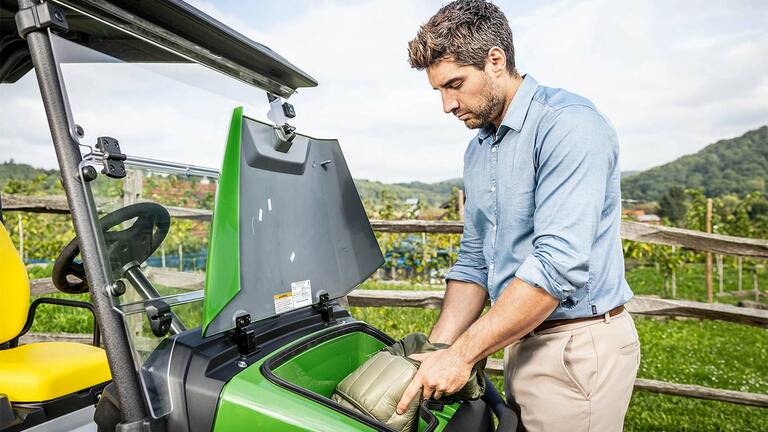 Persona ievieto polsterētu somu zaļa John Deere Gator GS priekšējā glabāšanas nodalījumā, kas atrodas ārā netālu no koka žoga un vīna dārza.