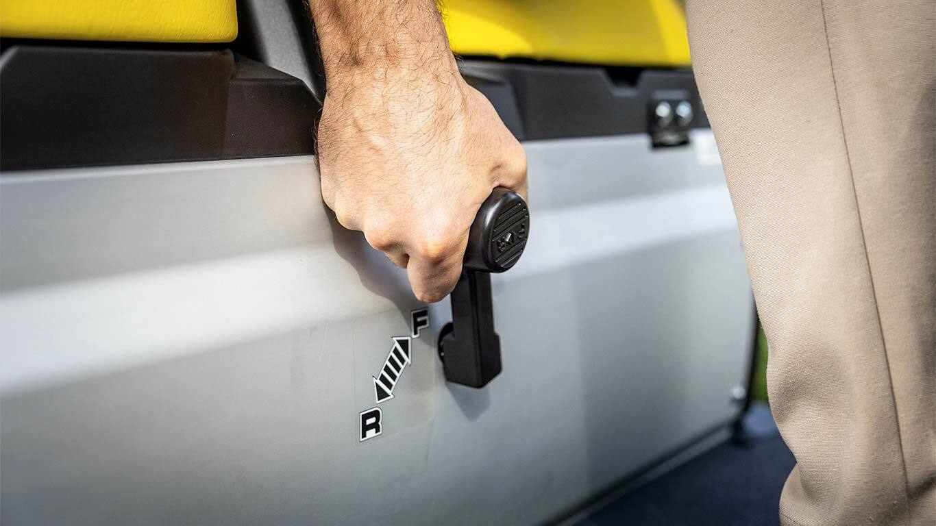 Tuvplāns ar roku, kas darbina John Deere Gator GS pārnesumu pārslēgšanas sviru, parādot turpgaitas un atpakaļgaitas stāvokļus blakus dzeltenajiem sēdekļiem.