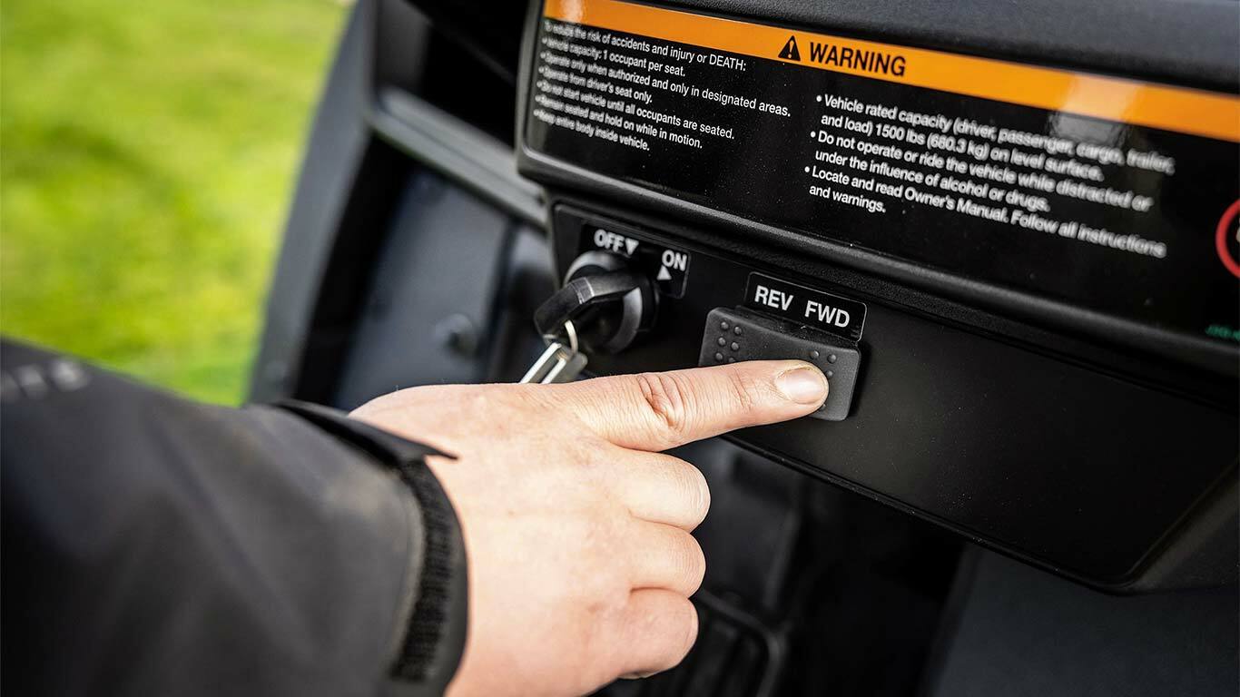 Pirksts, kas nospiež turpgaitas pārnesuma selektora pogu John Deere Gator GS elektriskā modeļa vadības panelī.