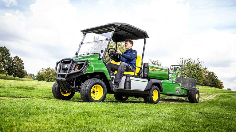 Zaļš John Deere Gator elektriskais universālais transportlīdzeklis, kas velk aprīkojumu pa pļavu ar daļēji mākoņainām debesīm fonā.