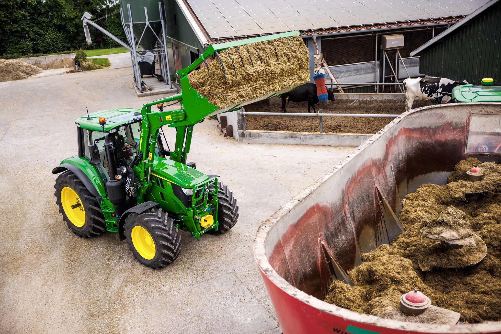 John Deere 6M sērijas traktors izmanto priekšējo iekrāvēju, lai iepildītu skābbarību maisītāja vagonā, saimniecības pagalmā.