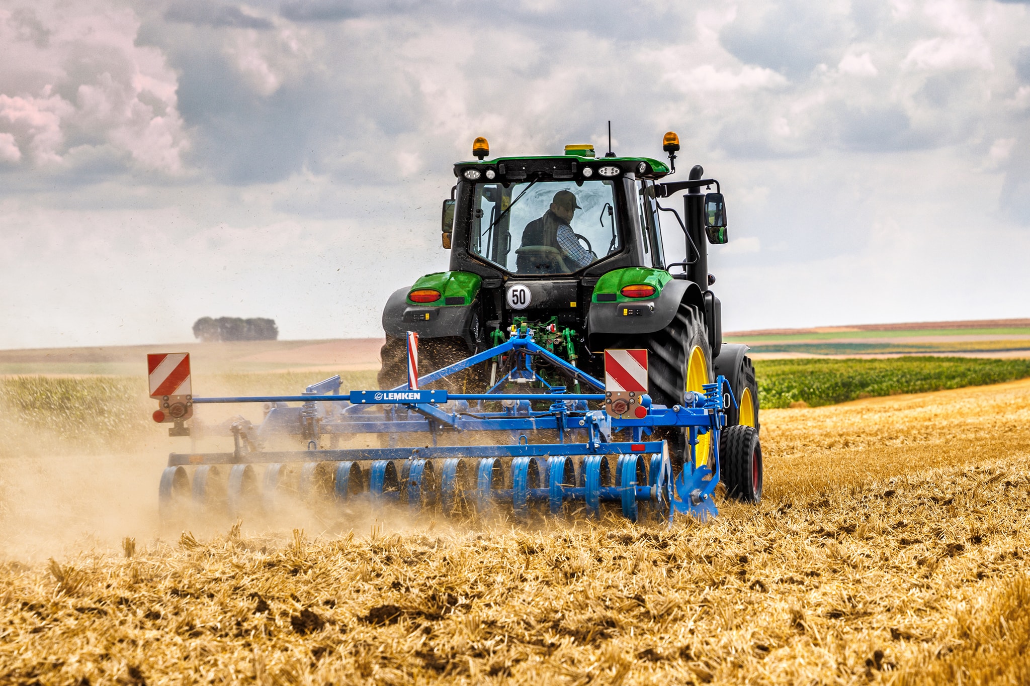 John Deere 6M sērijas traktors, kas kultivē jau apstrādātu lauku ar diska ecēšām, skats no aizmugures, uz putekļaina lauka.