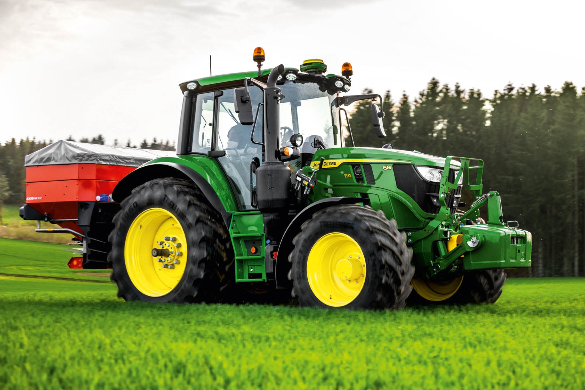 John Deere 6M sērijas traktors ar aizmugurē uzstādītu izkliedētāju, kas darbojas zālē, mežmalas tuvumā.