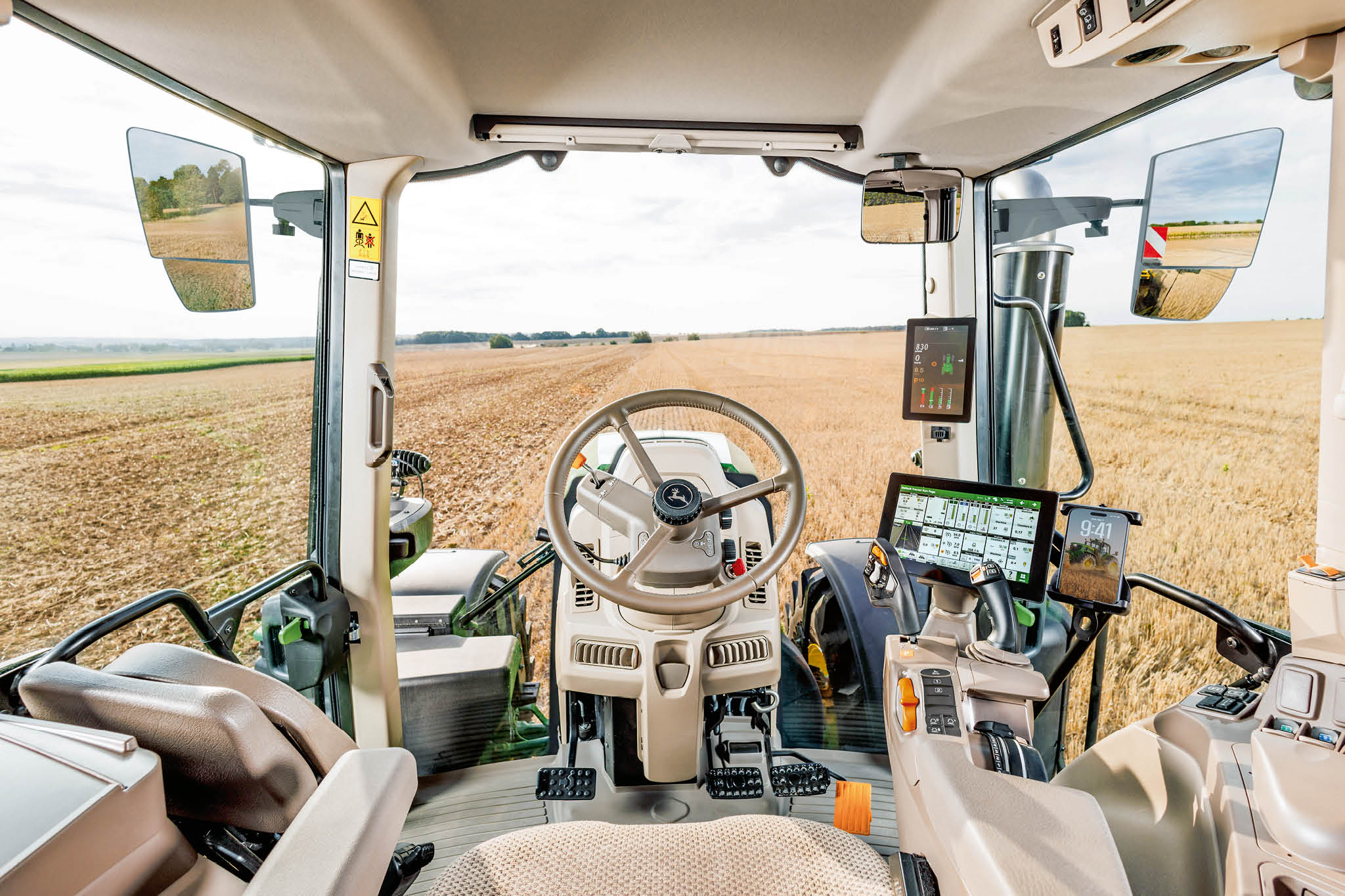 John Deere traktora kabīnes iekšpuse ar stūri, digitālajiem vadības displejiem un skatu uz lauku caur vējstiklu lauksaimniecības darbu laikā.