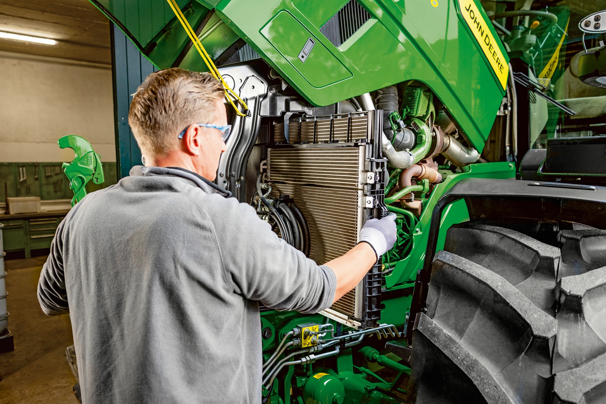 Apkopes tehniķis veic apkopi John Deere 6M sērijas traktora dzinēja nodalījumā.