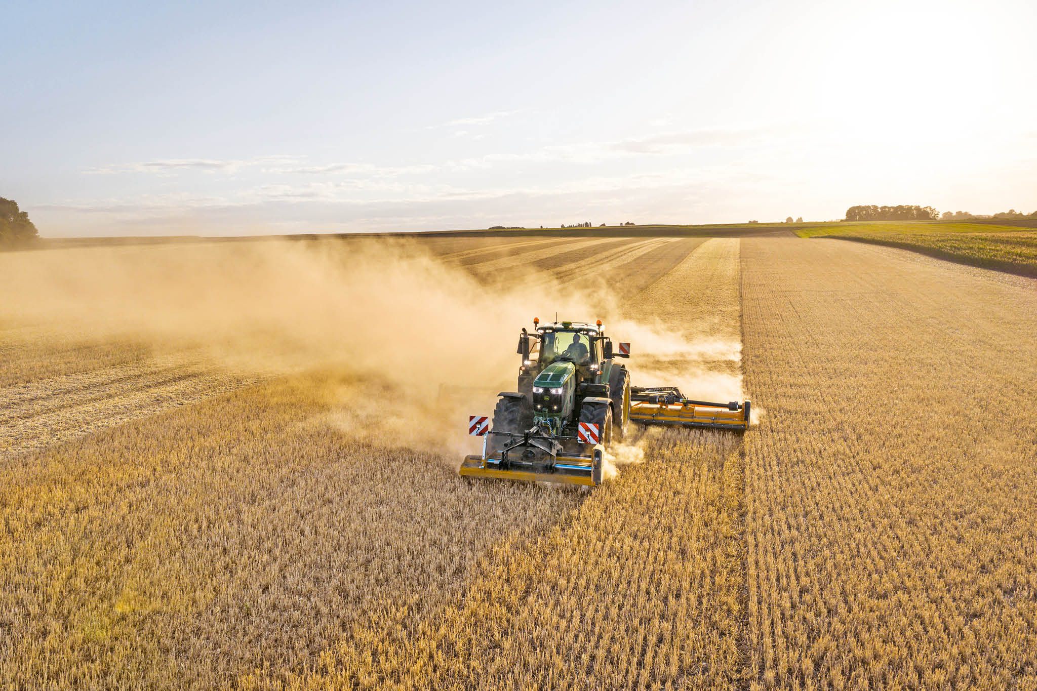 John Deere 6M sērijas traktors velk vircas cisternu pa lauku ceļu, kas robežojas ar zāli un laukiem.