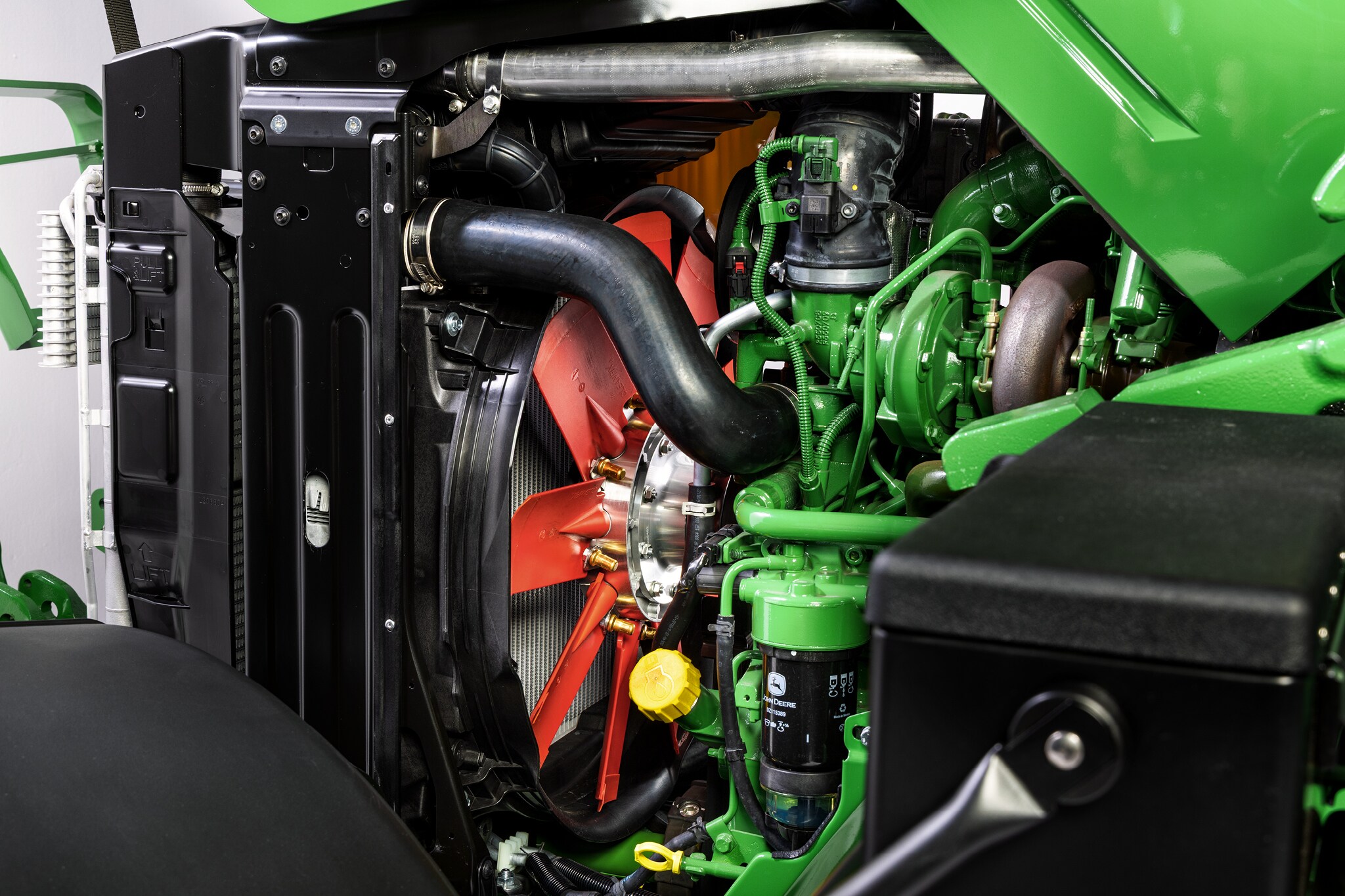 John Deere 6M sērijas dzinēja nodalījuma skats no augšas, kurā izcelta dzesēšanas sistēma un dzinēja komponenti.
