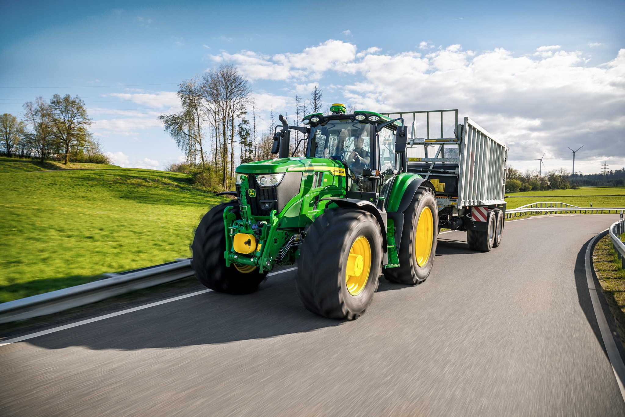 John Deere 6M sērijas traktors, kas pārvadā piekabi ar augstām malām, pa asfaltētu lauka ceļu.