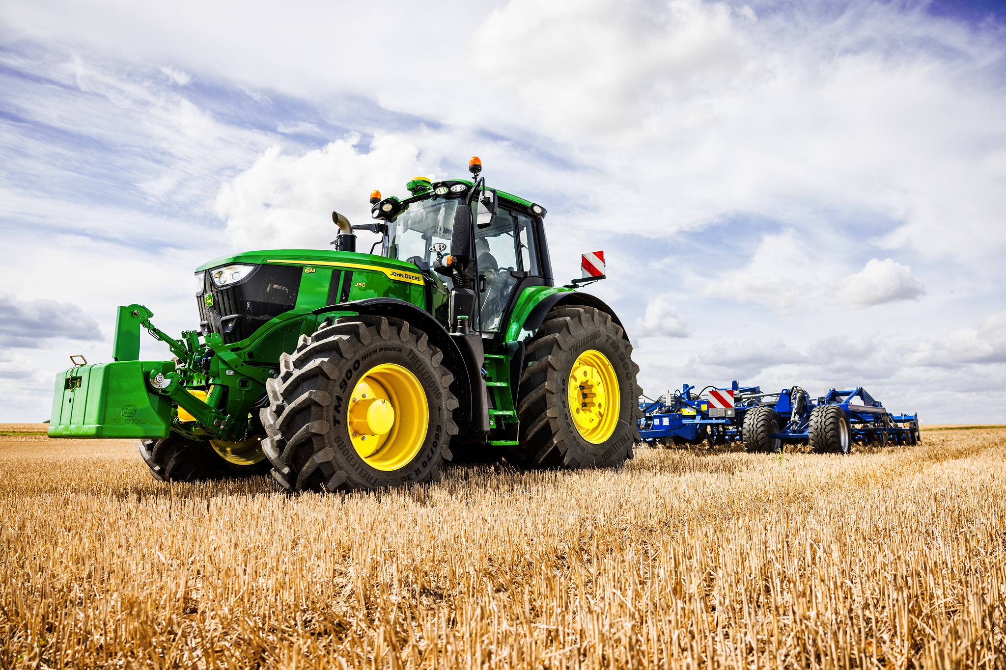 John Deere 6M sērijas traktors velk smagu aršanas agregātu pa stublāju lauku.