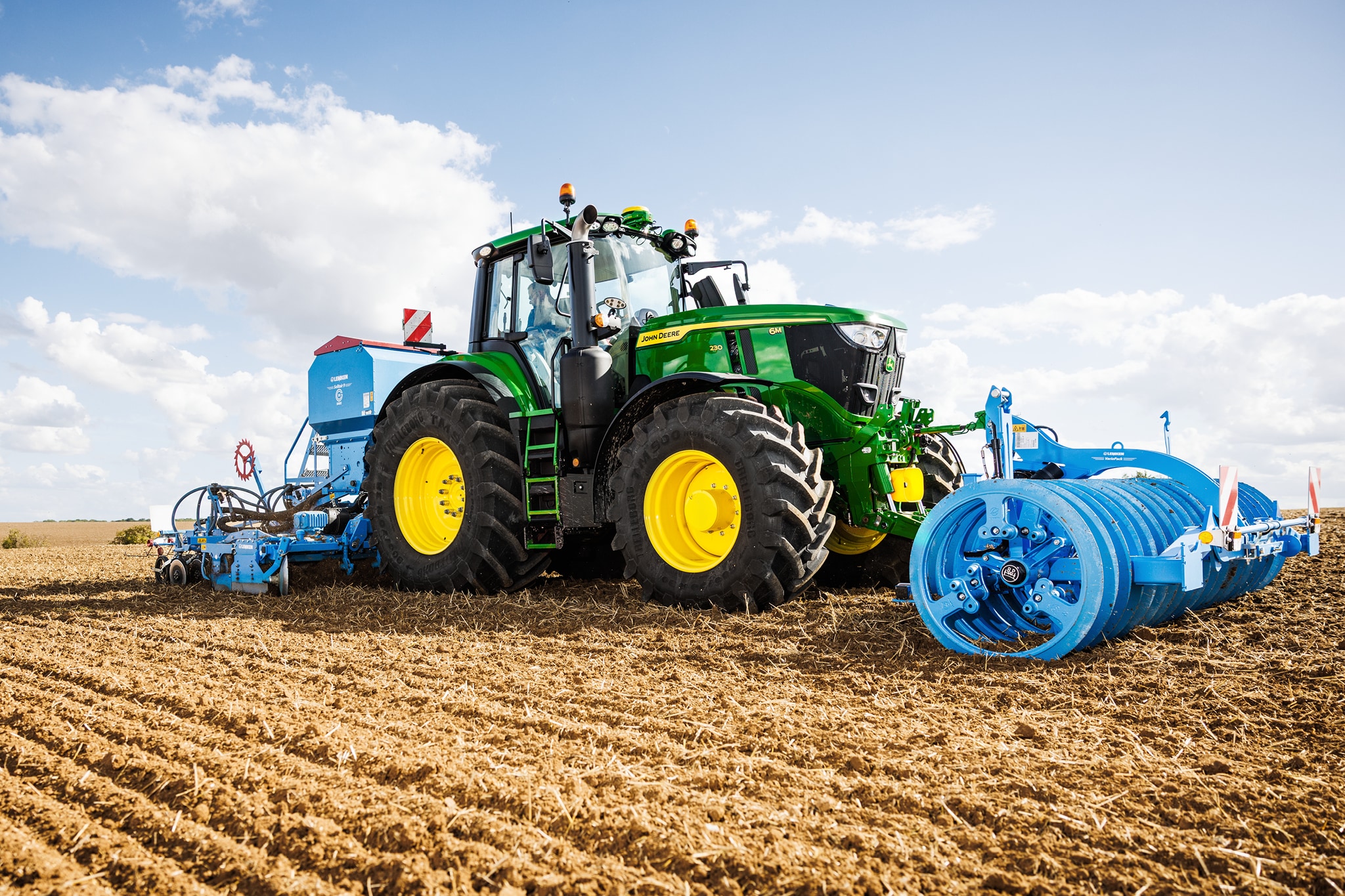 John Deere 6M sērijas traktors, kas kultivē lauku ar pievienotu lielu zilu veltņu agregātu.
