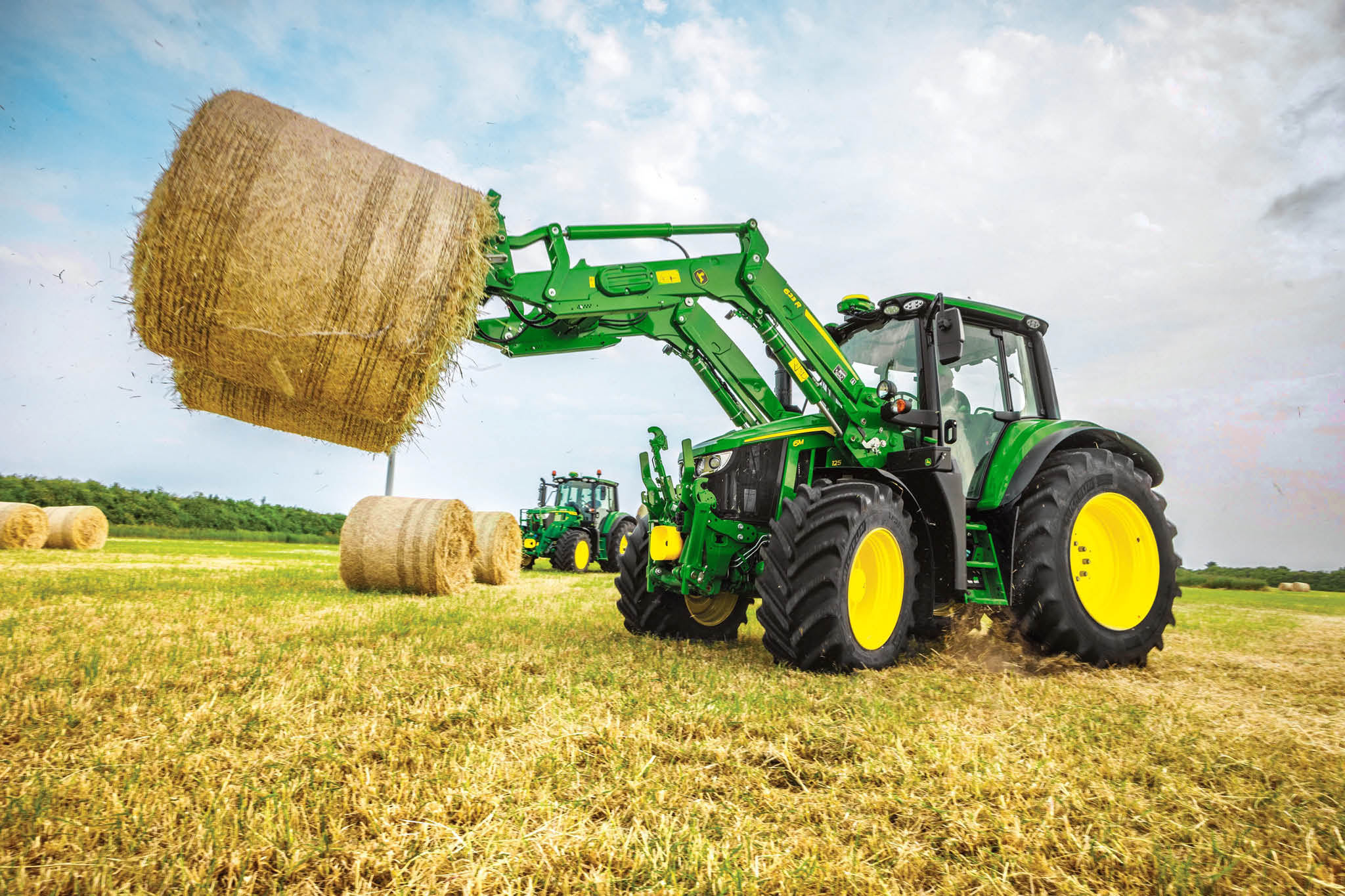 John Deere 6M sērijas traktors ceļ lielu apaļu siena ķīpu ar priekšējo iekrāvēju uz lauka.