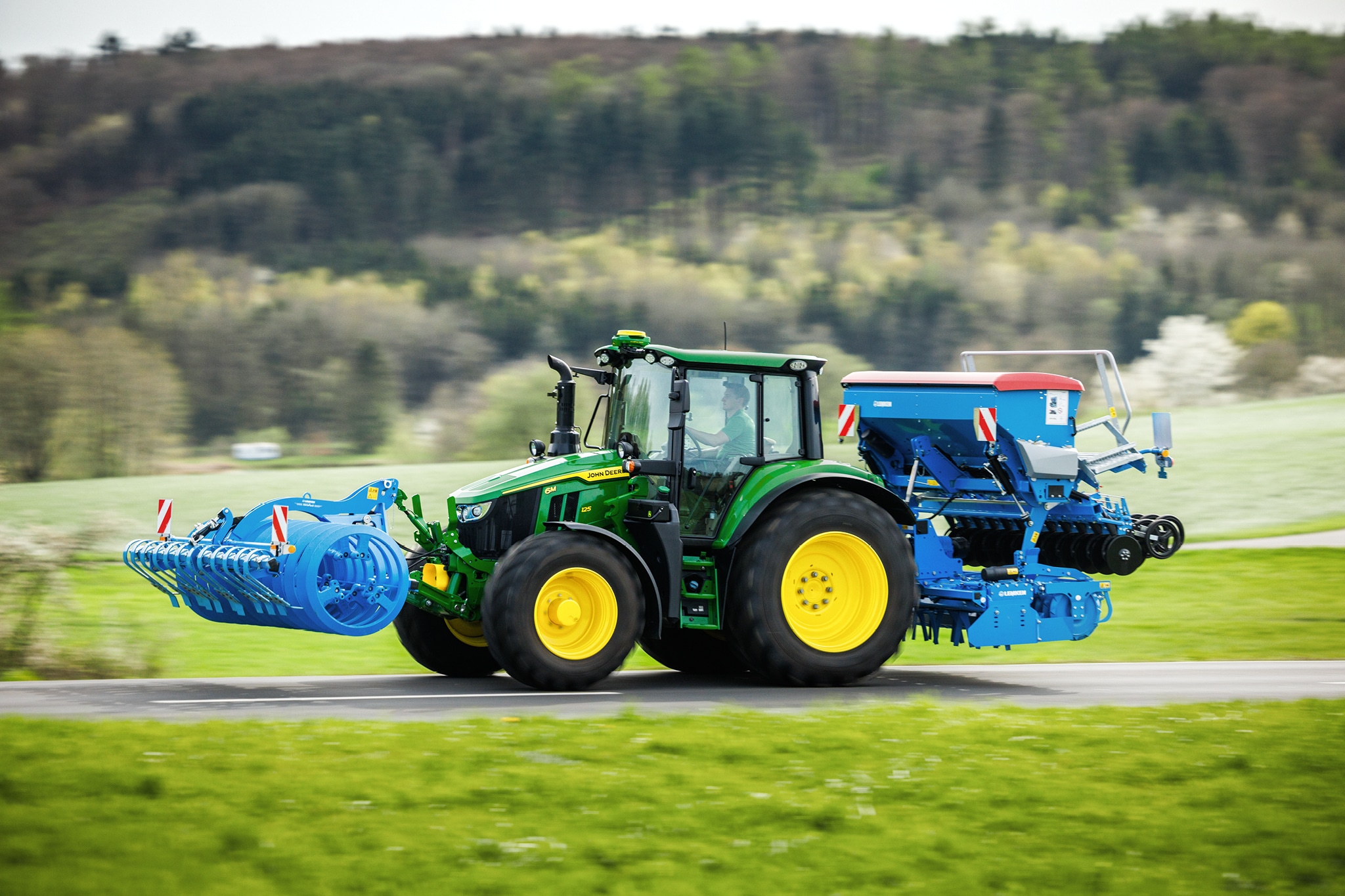 John Deere 6M sērijas traktors, kas transportē priekšā uzstādītu agregātu un aizmugurējo sējmašīnu pa lauku ceļu.