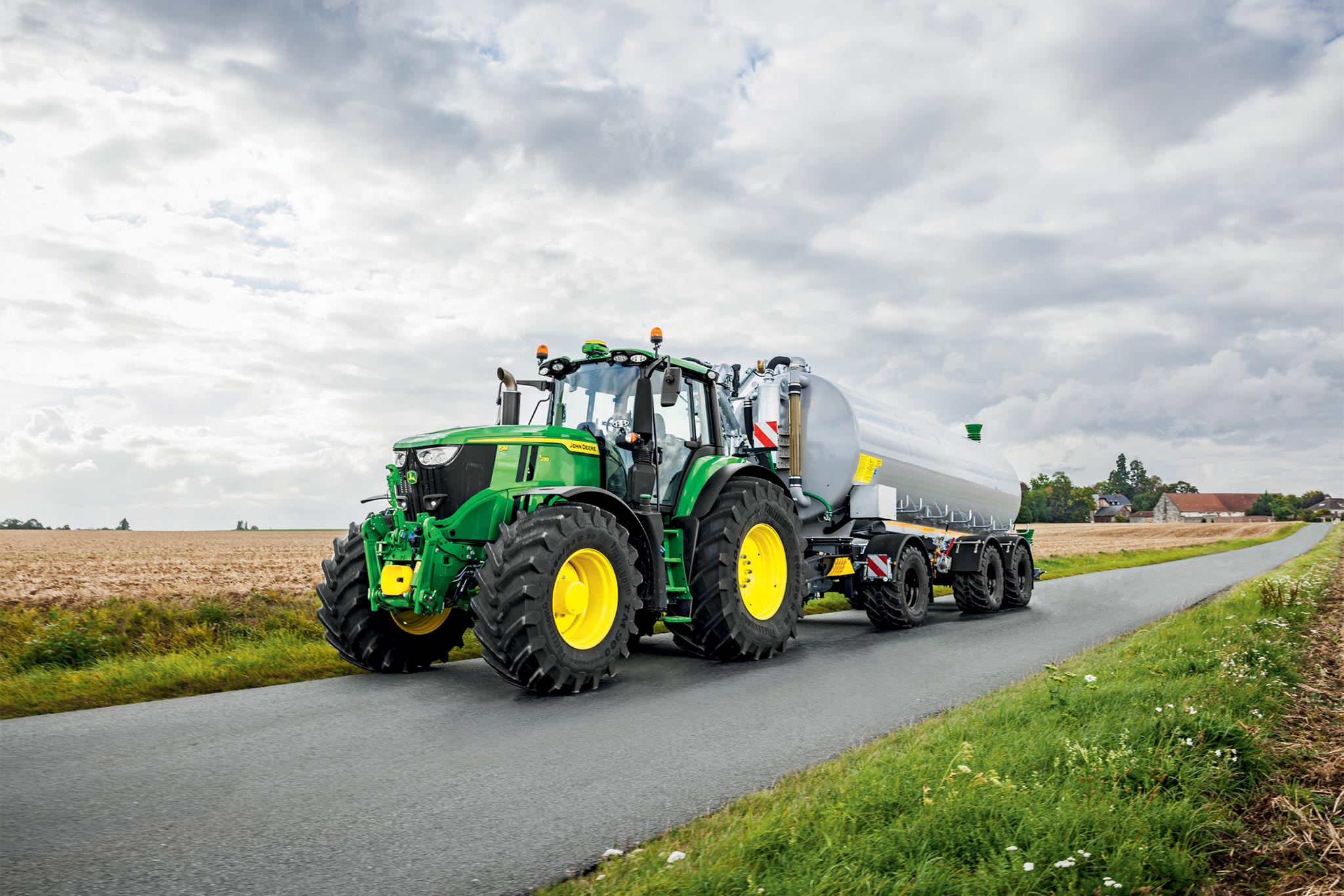 John Deere 6M sērijas traktors, kas strādā uz jau apstrādāta, putekļaina lauka ar platiem kultivatoriem, skatoties no augšas.