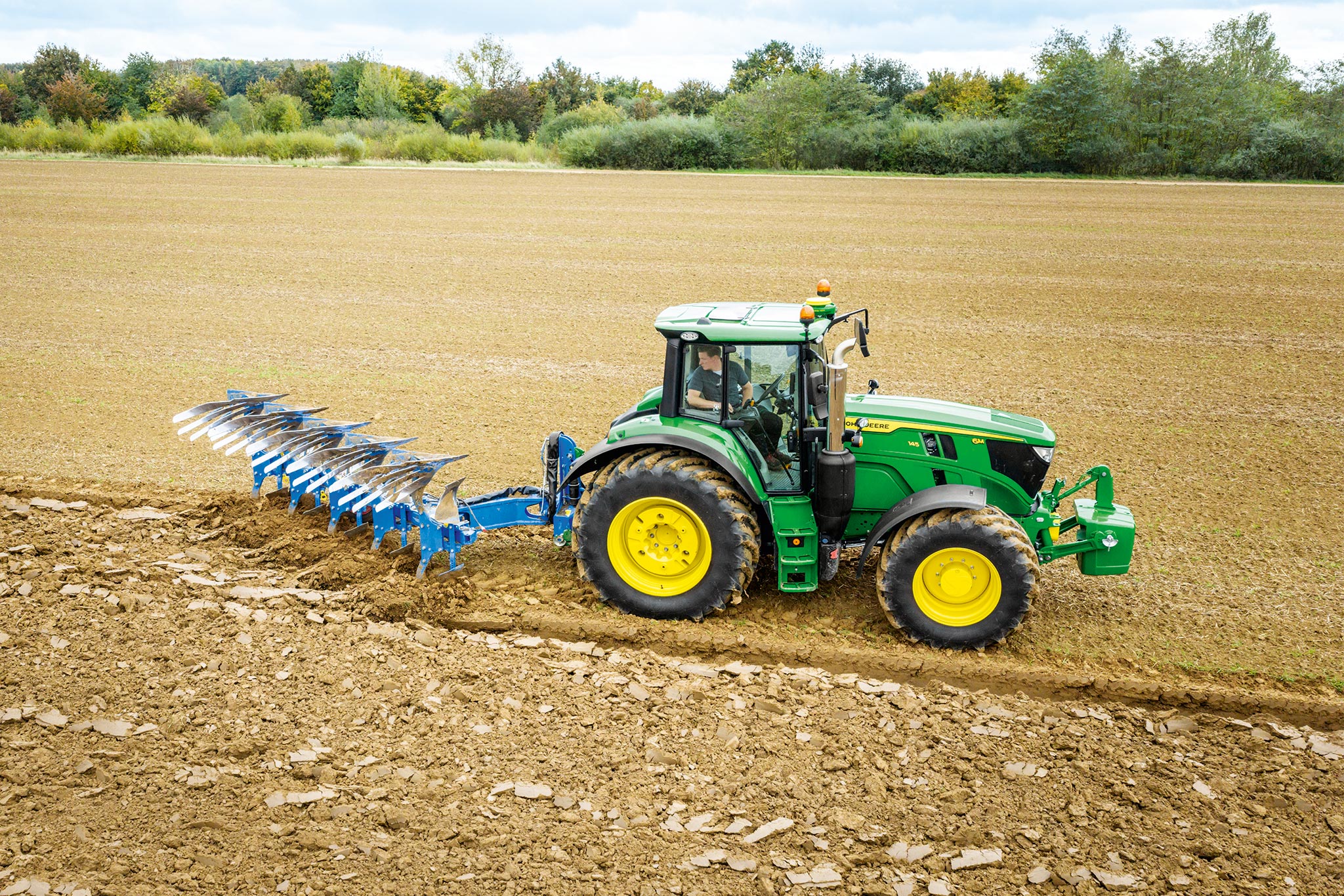 John Deere 6M sērijas traktors strādā uz aparta lauka ar aizmugurē uzstādītu aršanas agregātu.