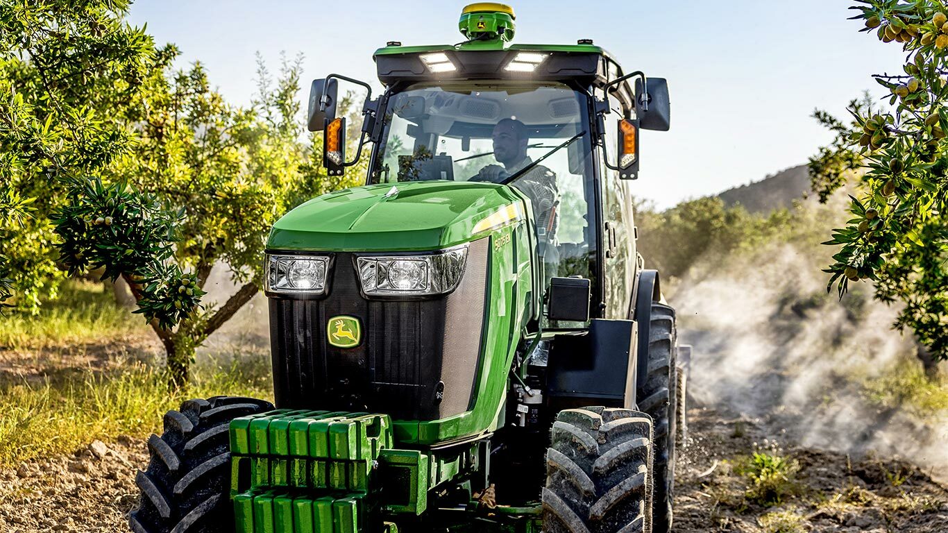 Zaļš un dzeltens John Deere 5075EN traktors, kas brauc cauri augļu dārzam pa zemes ceļu, ko ieskauj augļu koki un saules gaisma.