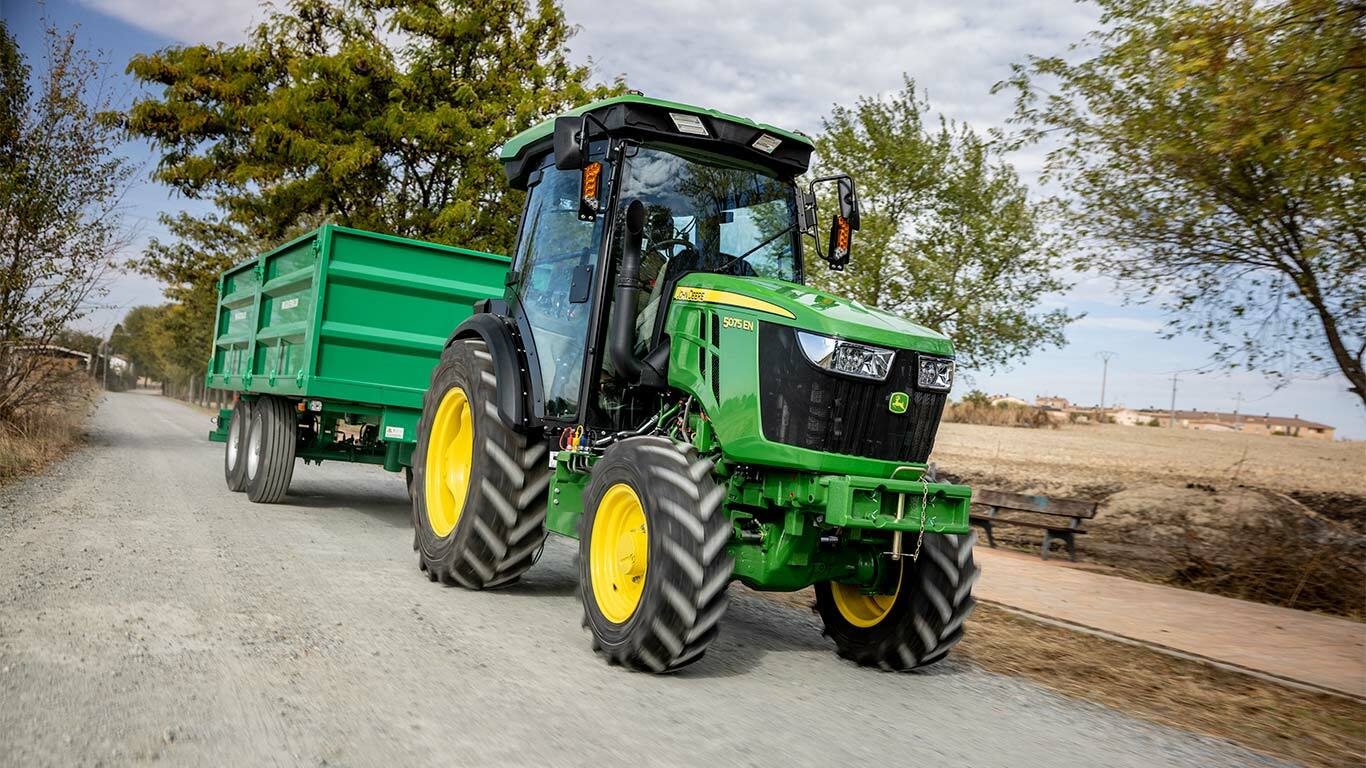 John Deere 5075EN traktors velk zaļu piekabi pa lauku ceļu ar kokiem fonā.
