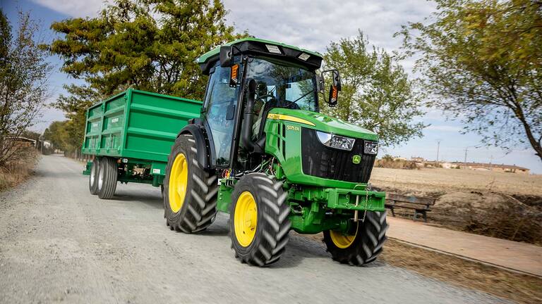 John Deere 5075EN traktors velk zaļu piekabi pa lauku ceļu ar kokiem fonā.