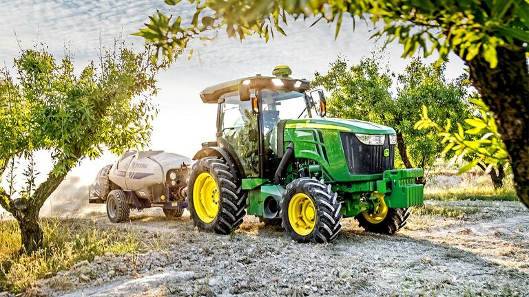 Zaļš un dzeltens John Deere 5090EN traktors velk smidzinātāju cauri augļu dārzam saulainā dienā.
