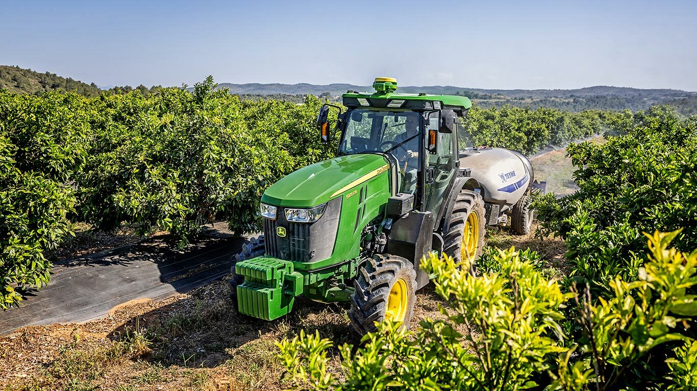 Zaļš un dzeltens John Deere 5105EN traktors velk smidzinātāju bagātīgā augļu dārzā ar paugurainiem pakalniem fonā.