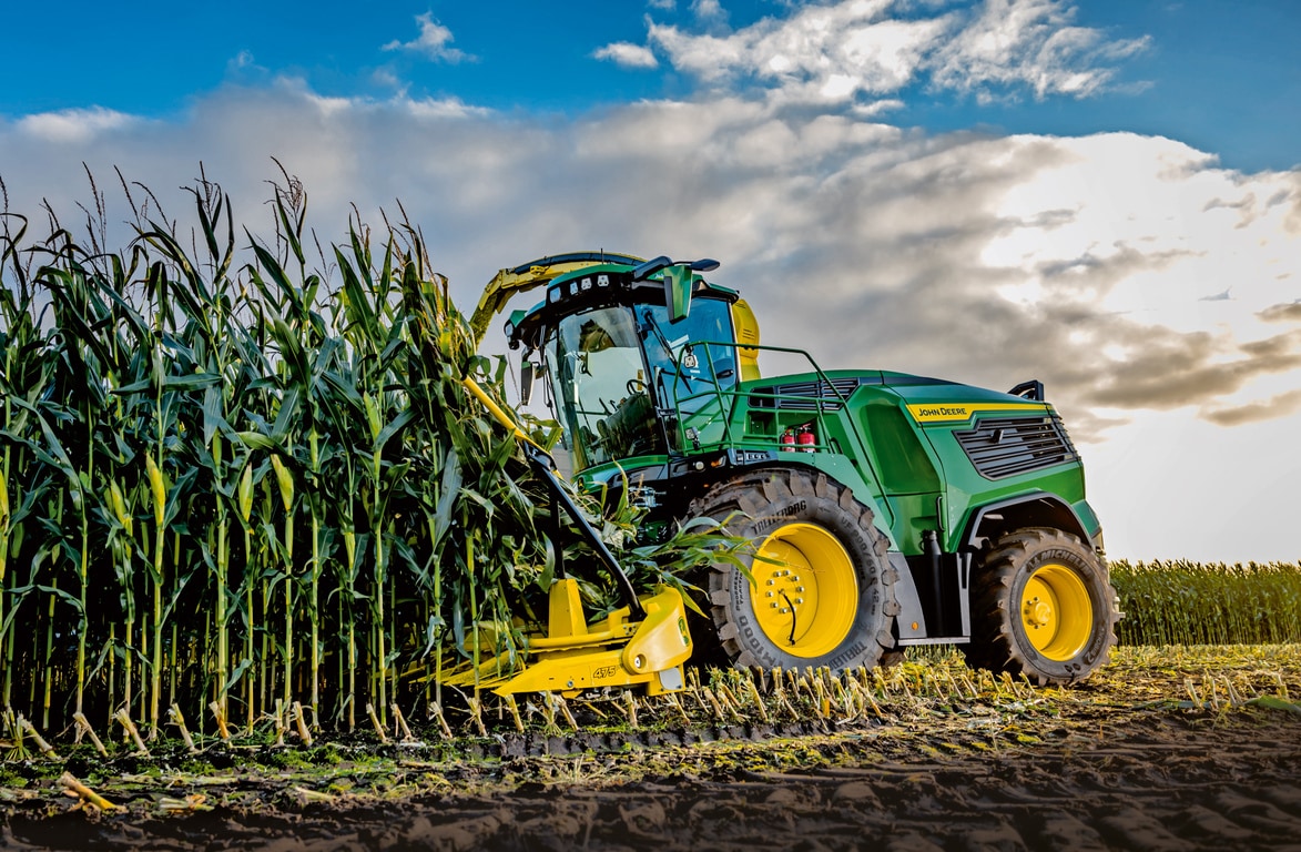 John Deere lopbarības kombains novāc kukurūzu uz lauka zem zilām debesīm ar mākoņiem.