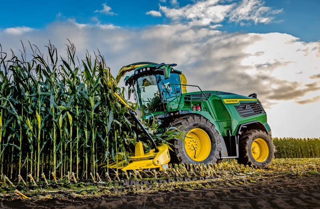 John Deere lopbarības kombains novāc kukurūzu uz lauka zem zilām debesīm ar mākoņiem.