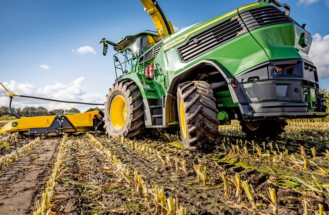 John Deere lopbarības kombains strādā novāktā kukurūzas laukā, parādot mašīnas lielās riepas un pļaušanas mehānismu uz skaidru zilu debesu fona.
