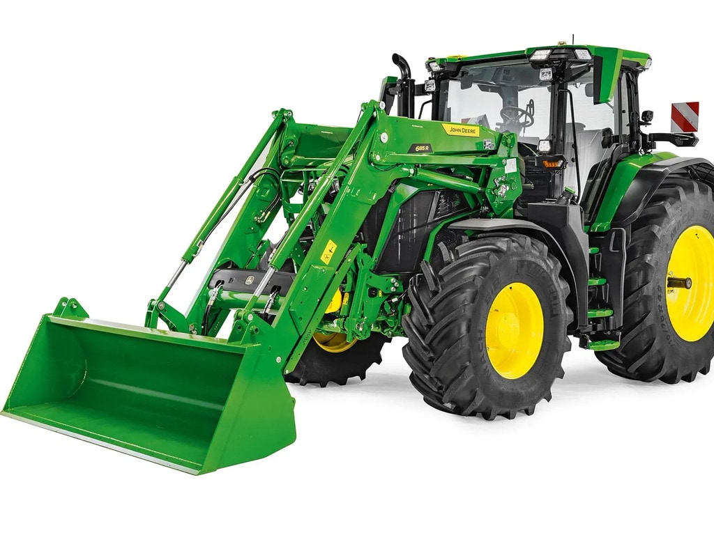 John Deere traktors ar pagarinātu priekšējo iekrāvēju un kausu, profesionāls fotoattēls, kas izceļ iekrāvēja sniedzamību un pacelšanas augstumu.