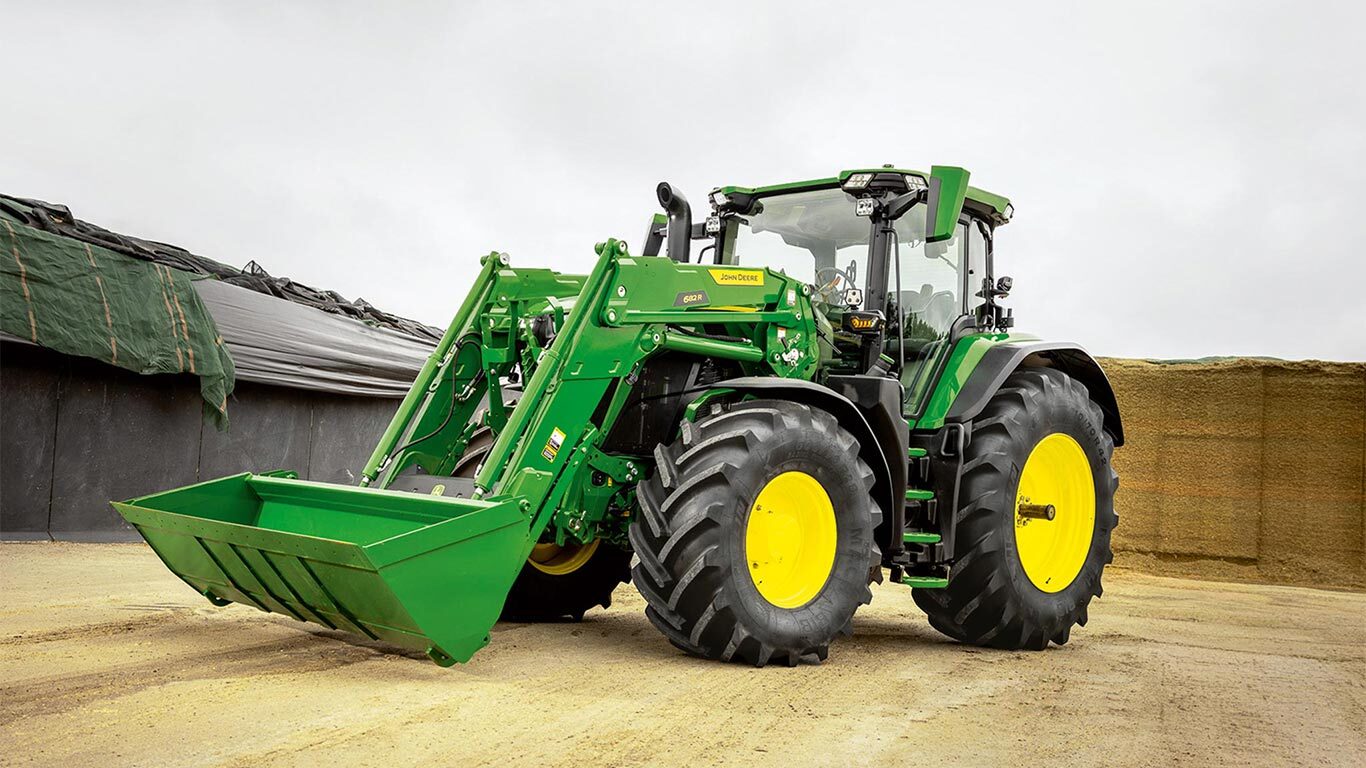 John Deere traktors ar priekšējo iekrāvēju novietots pagalmā netālu no skābbarības uzglabāšanas vietas.