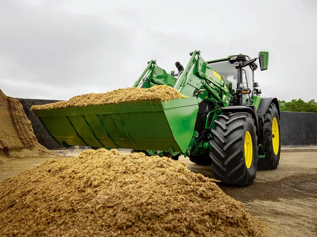 John Deere traktors ar priekšējo iekrāvēju pārvadā sasmalcinātu skābbarību lauksaimnieciskās uzglabāšanas zonā.