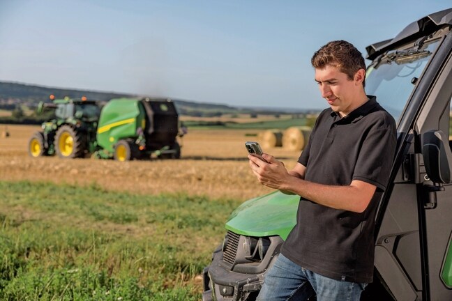Persona, kas pārbauda ruļļu dokumentācijas datus mobilajā tālrunī, fonā redzama John Deere VR prese