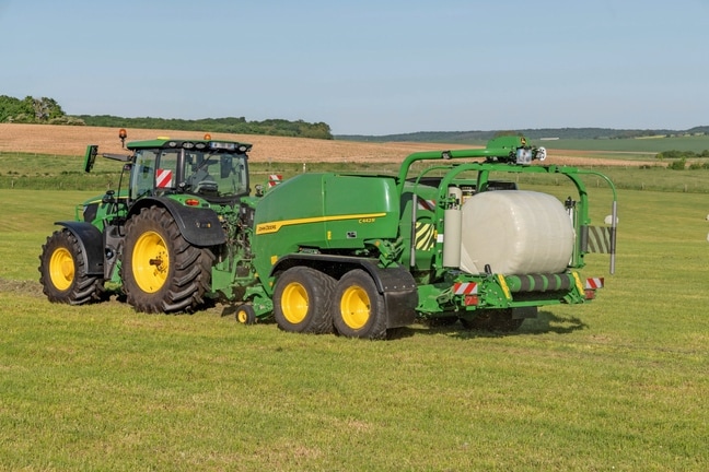 John Deere C442R ietīšanas ruļļu prese, ko velk John Deere 6R 185, veidojot liela blīvuma skābbarības ruļļus